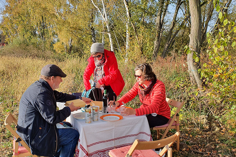 Auf einer Herbstwiese wird ein Campingtisch mit Tellern, Gläser und einer Flasche Rotwein gedeckt. In der beigen Assiette ist der Gänsebraten.